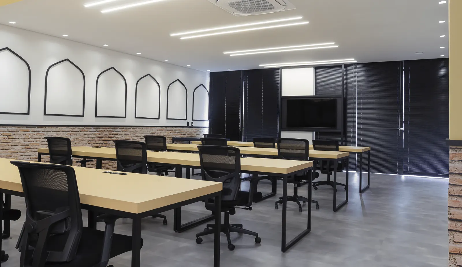 Sala de treinamento com mesas bege em formato escolar e TV na parede